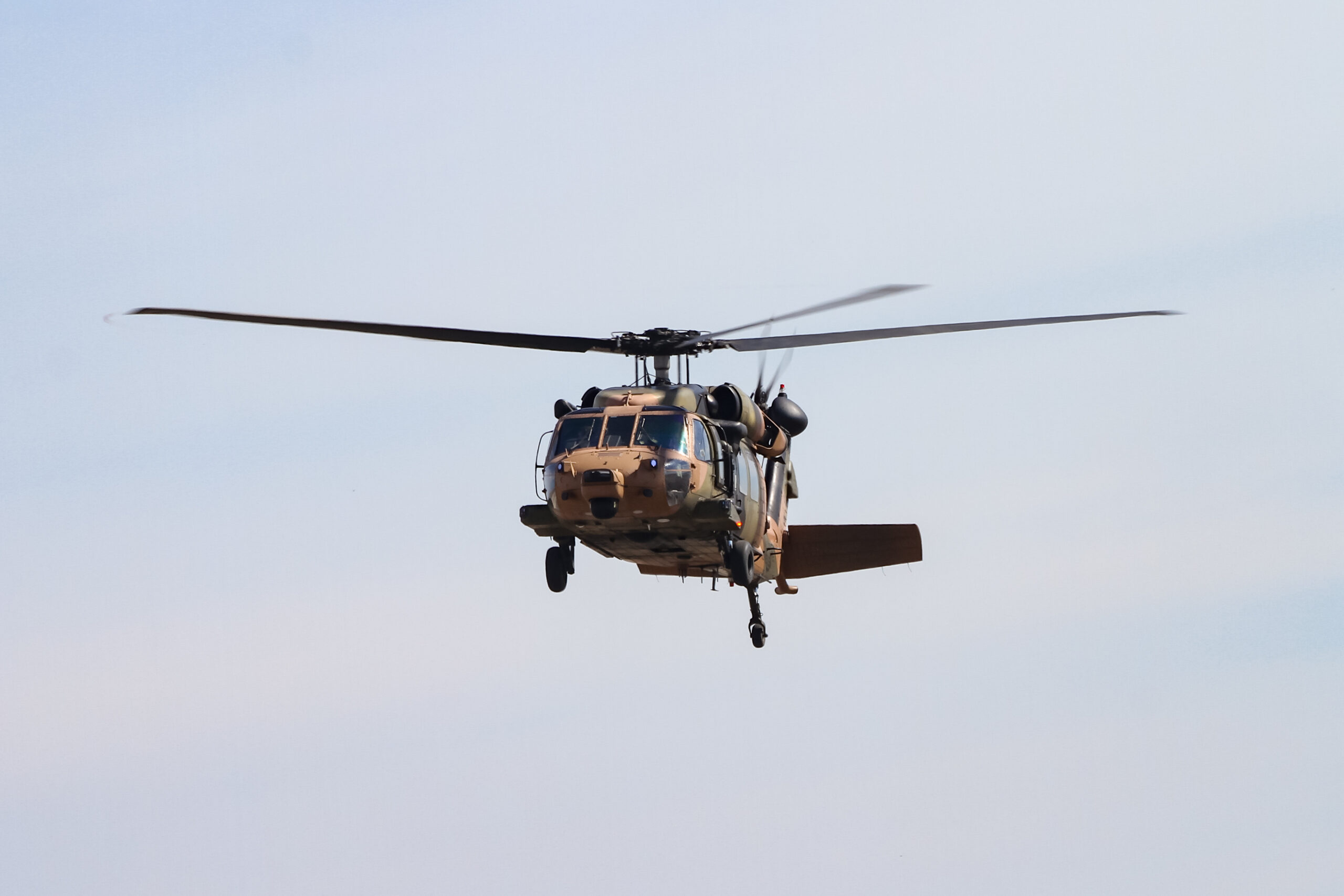 A Black Hawk military helicopter in flight against a clear blue sky. The helicopter features a camouflage paint scheme and is captured mid-air with its rotors in motion. The scene highlights the aircraft's design and operational state.