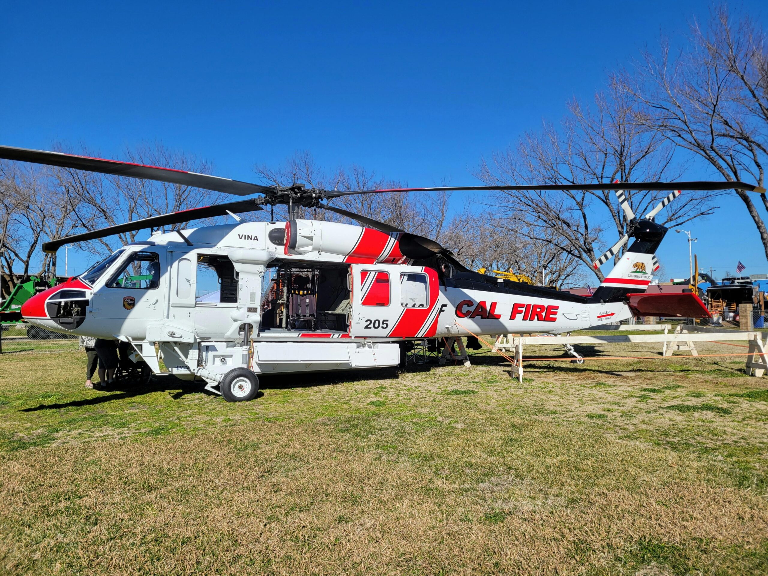 A CAL FIRE helicopter parked on a grassy field under a clear blue sky. The helicopter, marked with the number "205," features white and red markings with "CAL FIRE" prominently displayed on the side. Leafless trees and equipment can be seen in the background.